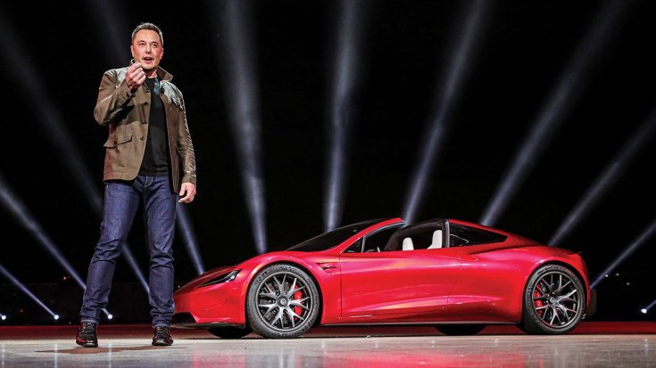 Elon Musk a színpadon magyaráz, háttérben új Tesla Roadsterrel