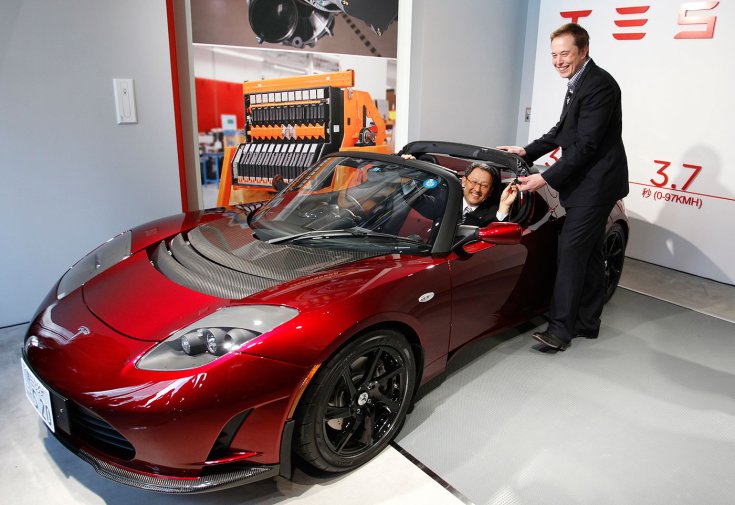 Akio Toyoda egy Tesla Roadsterben ülve, Elon Musk a kulcsokat adja át
