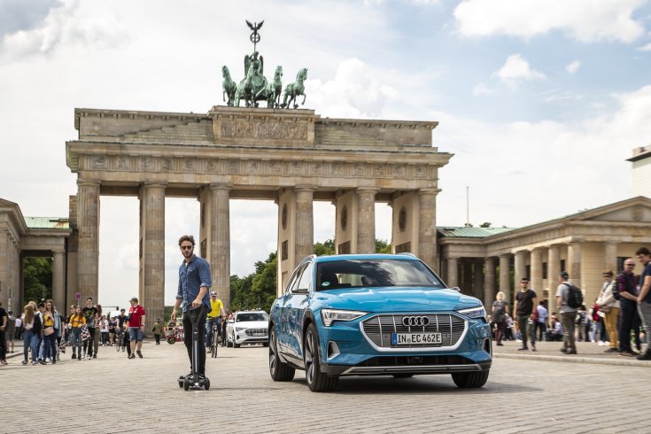 Egy férfi elektromos Audi rolleren egy Audi személygépjármű mellett