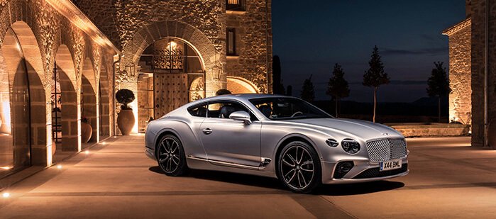 Egy esti kivilágításban tündöklő kastély előtt álló ezüst színű Bentley Continental GT
