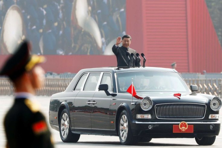 XI Jinping, a Kínai Kommunista Párt főtitkára integet egy fellobogózott Hongqi L5 limuzinból, az előtérben egy elmosódott kínai katona vigyázállásban