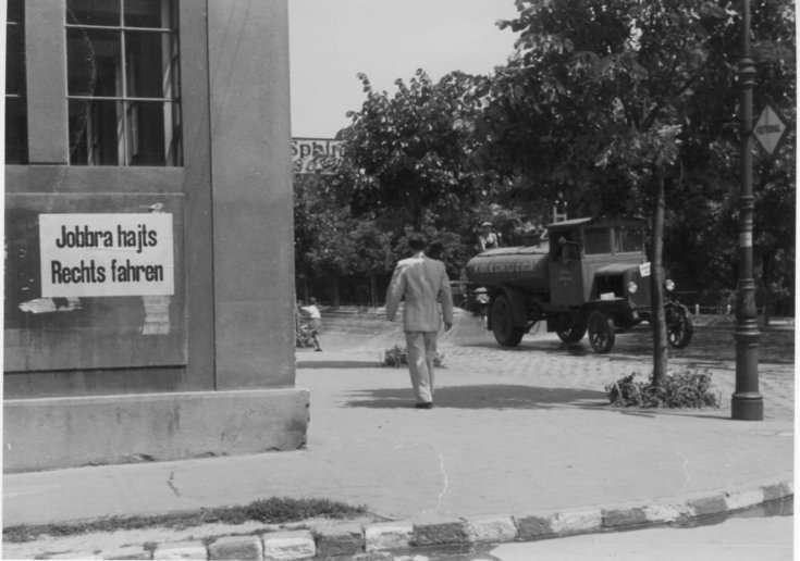 Egy „jobbra hajts” tábla egy ház oldalán, háttérben egy korabeli autó