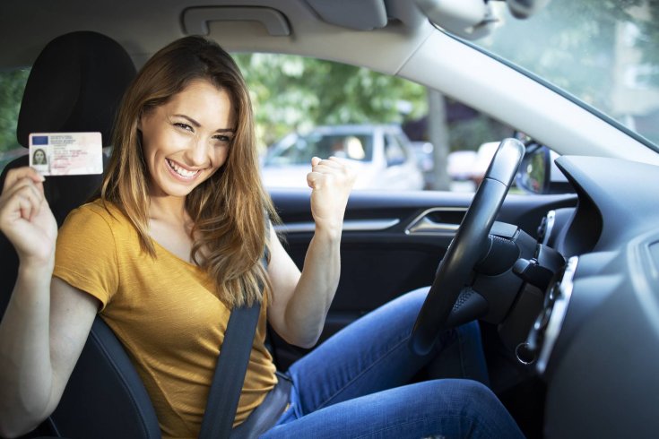 fotó egy autóban ülő boldog lányról, aki a jogosítványát mutatja felénk