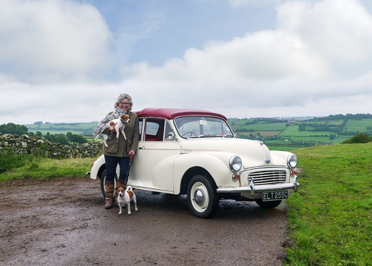 Morris Minor 1000, fehér kabrió, felhúzott vörös ponyvatető, elölnézet, oldalra döntve, jobbra, mellette egy nő 2 kutyával
